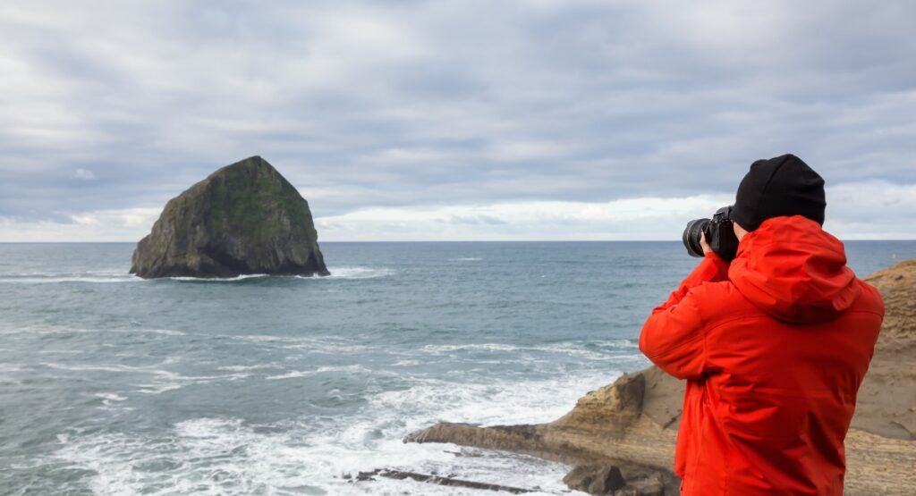 Photographer in a red jacket using a DSLR camera to capture the iconic Haystack Rock in the Pacific Ocean from a coastal viewpoint.