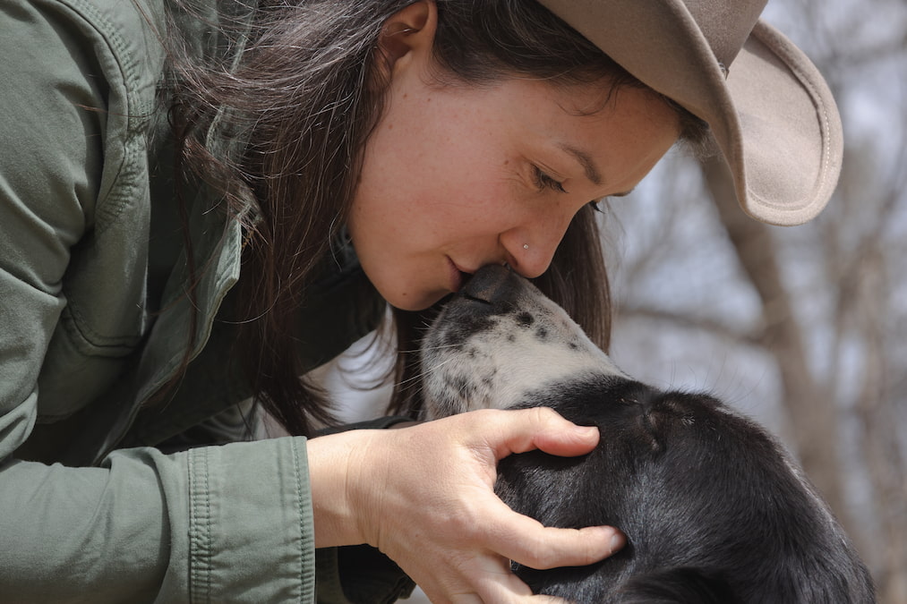 A woman in a cowboy hat gently nuzzles her black and white sheepdog, showing a close bond between human and working dog.